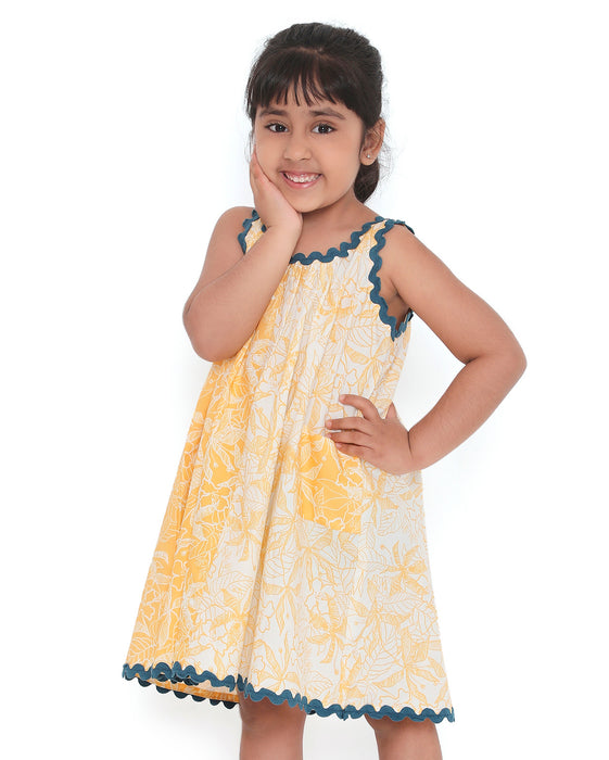 A young girl smiling and posing in a sleeveless yellow and white floral print dress with blue lace trim, standing with one hand on her cheek and the other on her hip