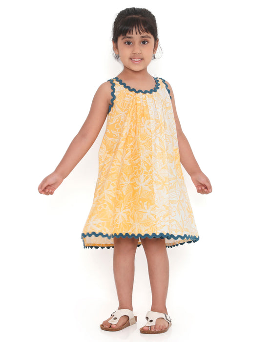 A young girl smiling and posing in a sleeveless yellow and white floral print dress with blue lace trim, standing with one hand on her cheek and the other on her hip