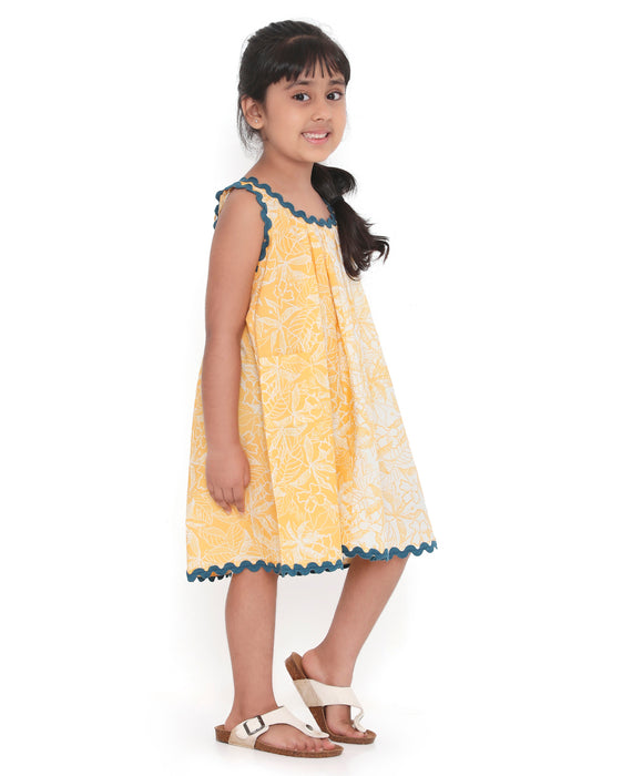 A young girl smiling and posing in a sleeveless yellow and white floral print dress with blue lace trim, standing with one hand on her cheek and the other on her hip