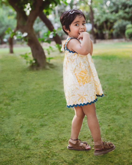 A young girl smiling and posing in a sleeveless yellow and white floral print dress with blue lace trim, standing with one hand on her cheek and the other on her hip