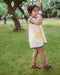 A young girl smiling and posing in a sleeveless yellow and white floral print dress with blue lace trim, standing with one hand on her cheek and the other on her hip